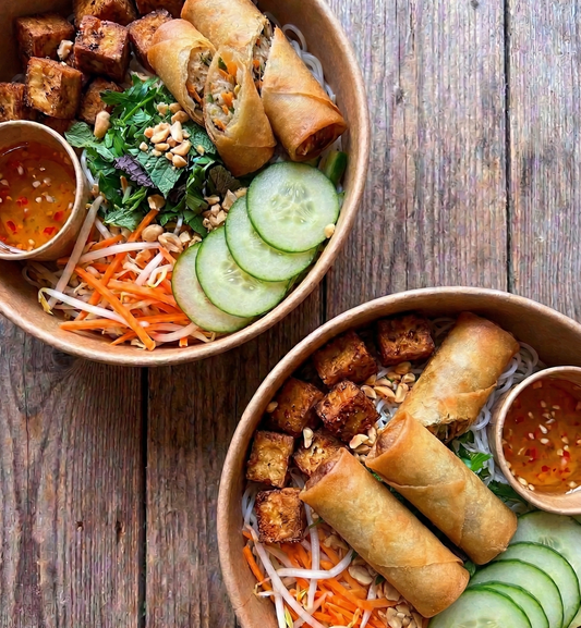 Two bowls of spring roll salad with vegetables and sauces on a wooden table.